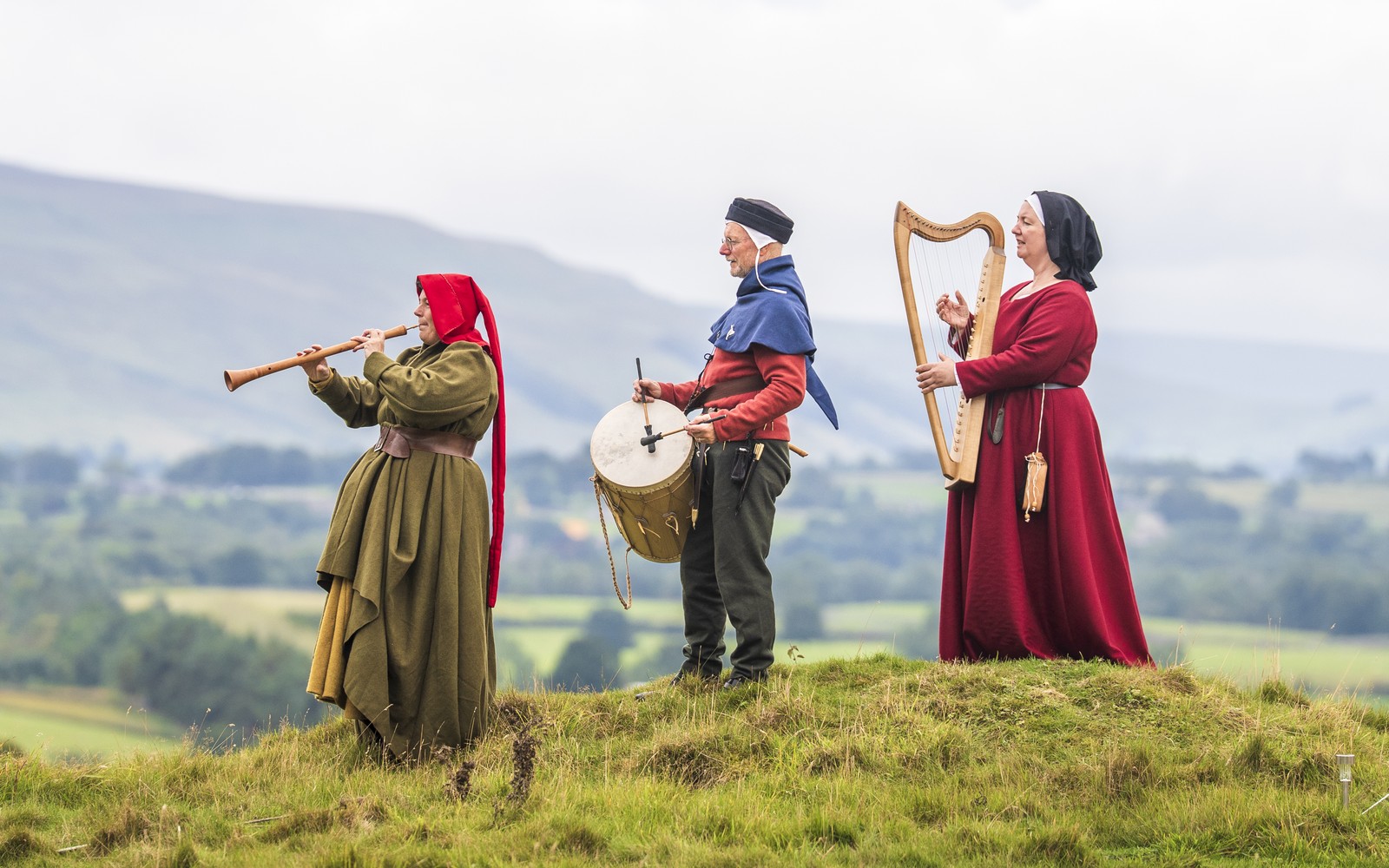 A trio of musicians wearing medieval clothing performs on a hilltop.