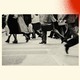 A black-and-white image shows people crossing the street. Some are carrying briefcases, and others are suit pants, long back skirts, and long trench coats.The photo is set against a background with three spray-painted circles. In the bottom-left corner is a blue one, in the bottom-right corner is a green one, and in the top-right corner is a red one.