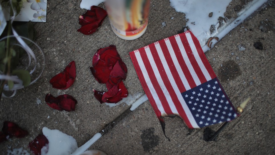 A candle, rose petals, and a partially intact American flag lie on a sidewalk