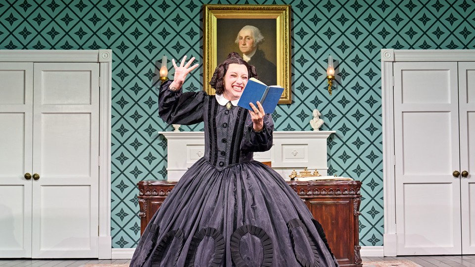 Photograph of a woman on stage, wearing an voluminous dress, reading from a book with a framed portrait of George Washington behind her