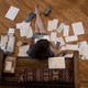 Woman sits on hardwood floor surrounded by scattered bills and paperwork