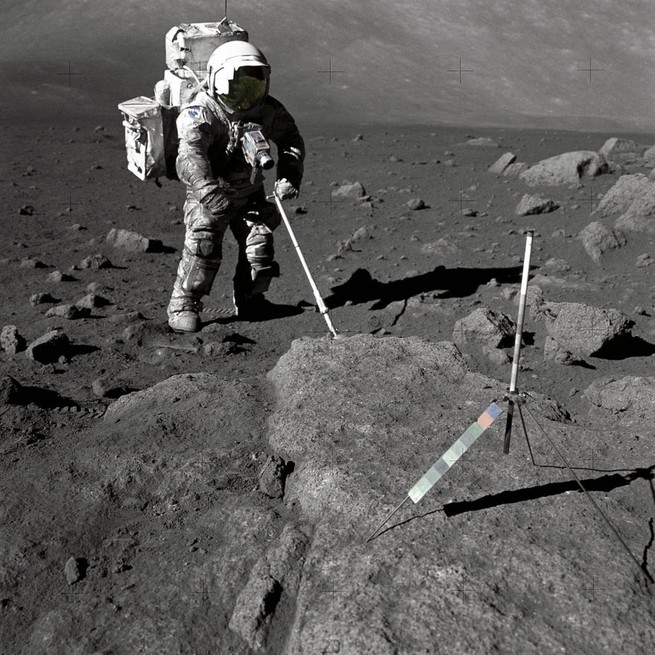 An astronaut uses a long instrument to probe rocks on the moon.