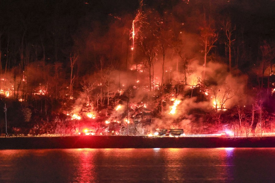A wildfire burns on a hillside, partly reflected in water.