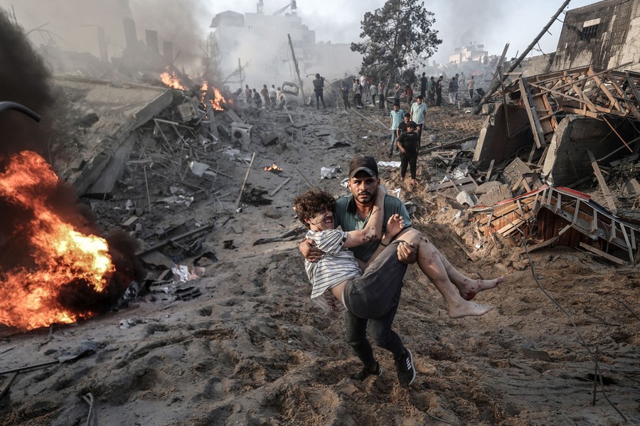 A person carries another, injured person over dirt and burning rubble, following an air strike.