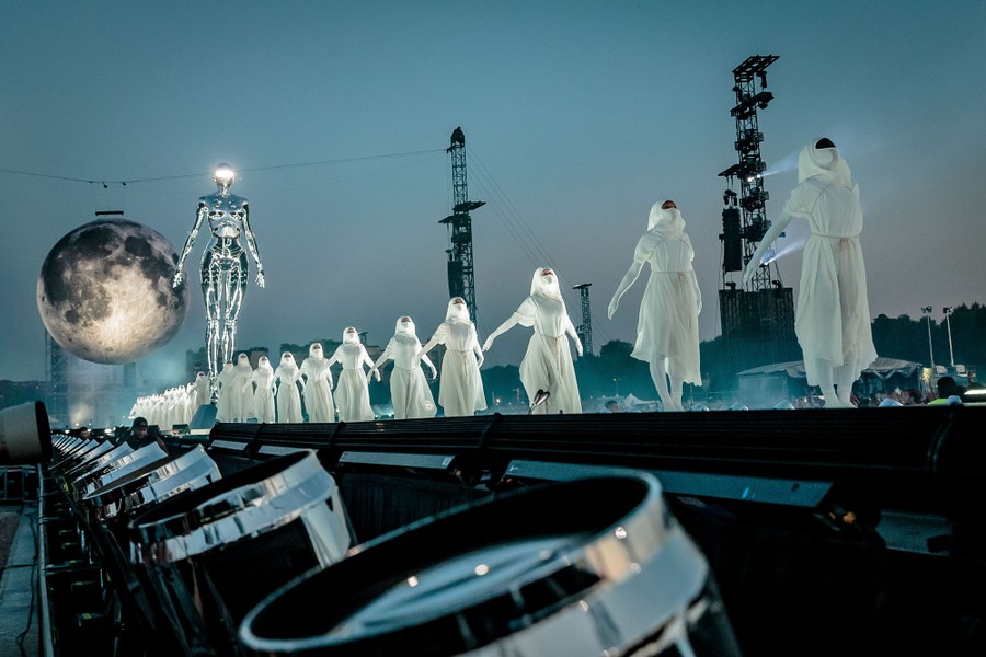 Performers stand in a line on a long stage in front of large sculptures of a robotic figure and the moon.
