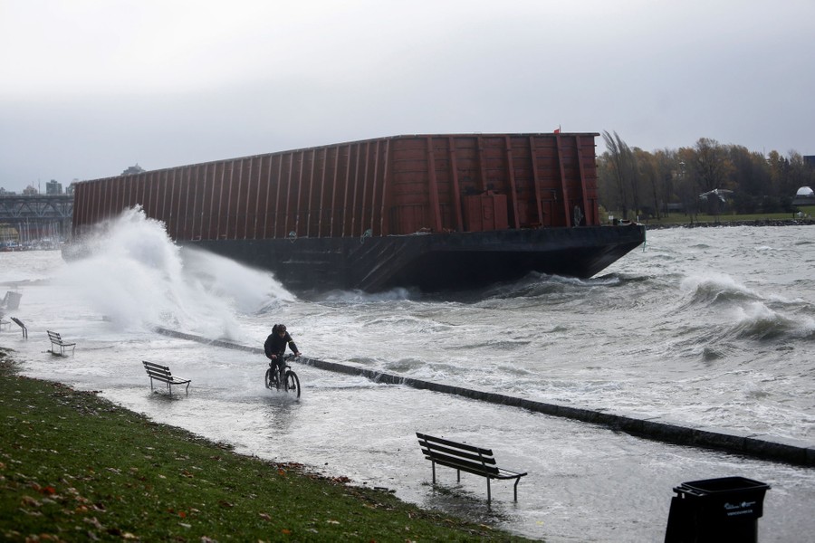 A person rides a bike on a flooded shoreline sidewalk, near a grounded barge, with waves splashing everything.