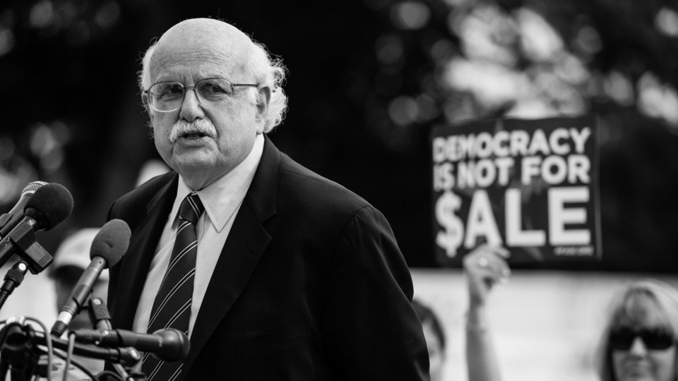 A black-and-white photo of Fred Wertheimer standing in front of a group of microphones; in the background a woman holds up a sign reading "Democracy Is Not for $ale."