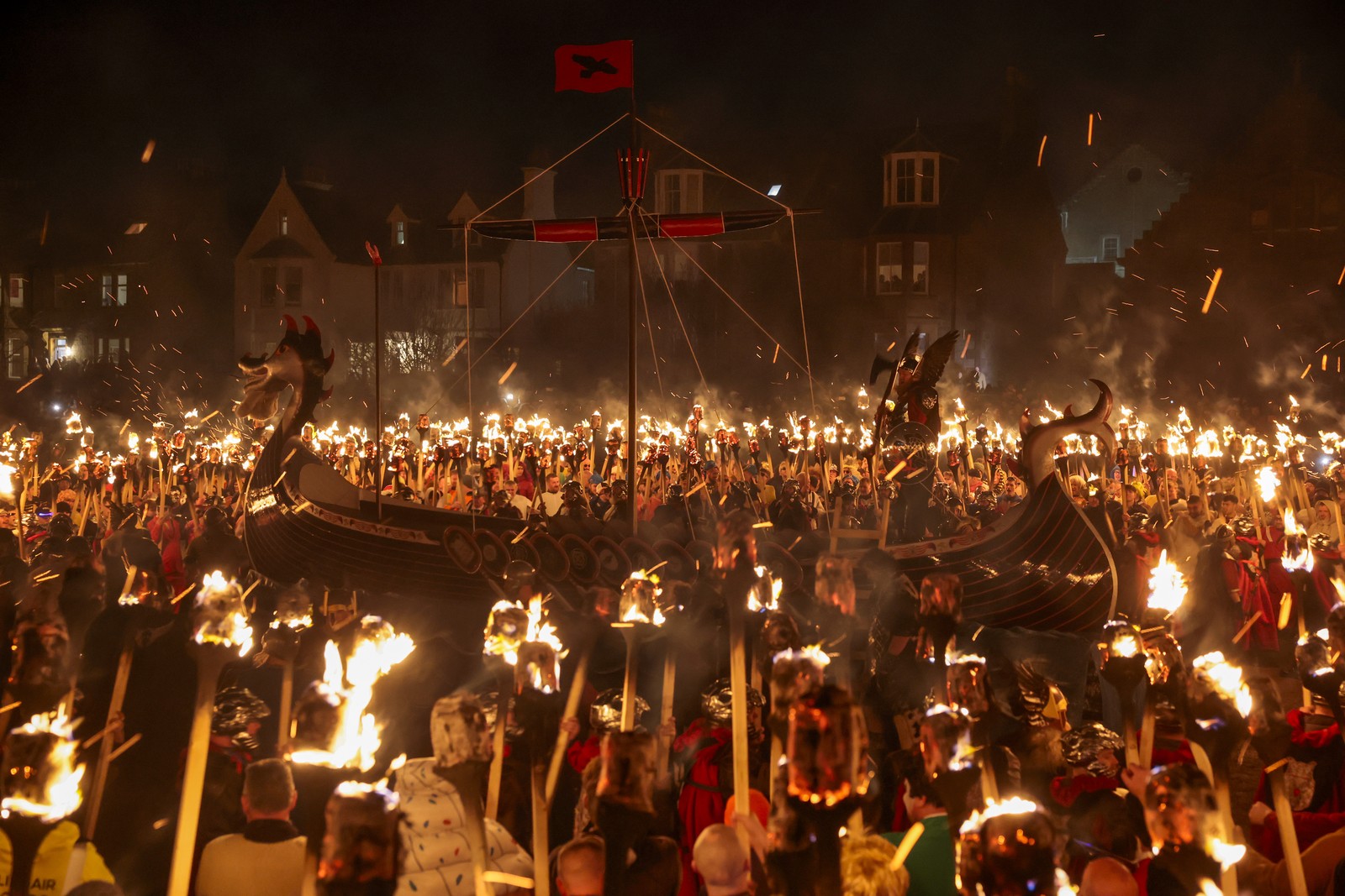A crowd of people holding flaming torches surrounds a replica Viking ship in a public square.