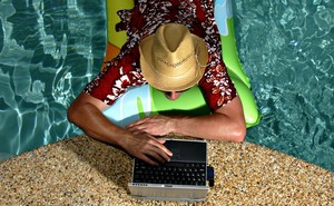 A photo of a man in a pool working on a laptop at the edge of the water.
