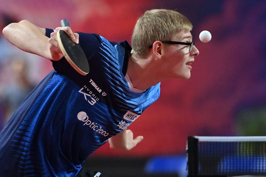 A table-tennis player leans forward to serve, a small white ball hanging just in front of their face.