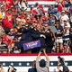A photograph of Donald Trump surrounded by Secret Service agents on a stage; people in the crowd looking on raise their arms and hold up their cameras.