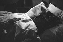 A woman  in pajamas reclines on a bed with her arm across her forehead.