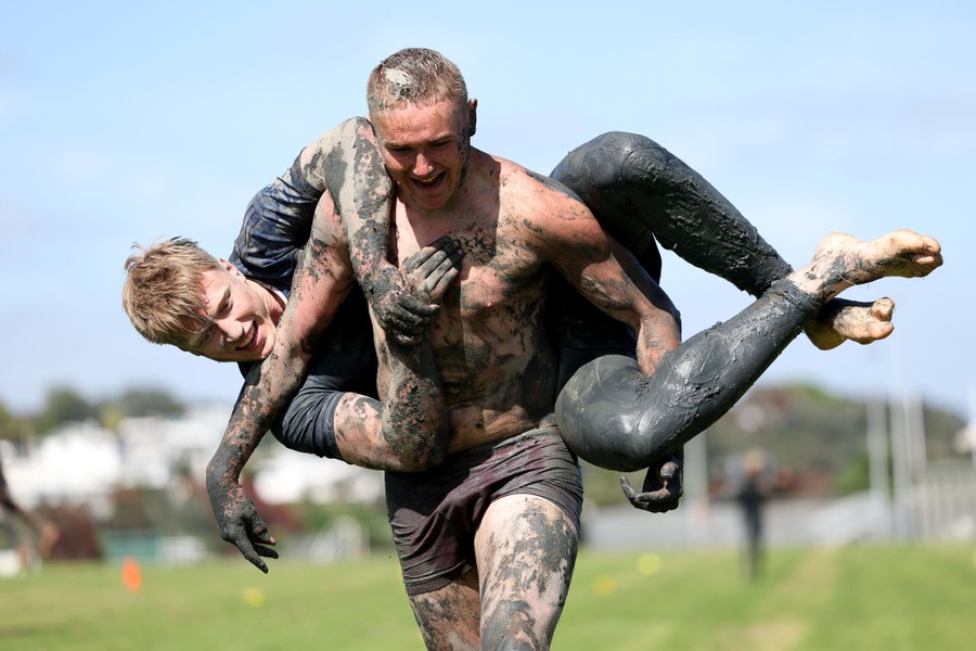 A person carries another person across their back—both are covered in mud.