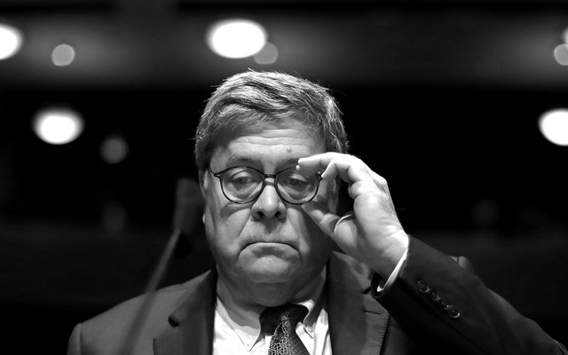 Shown in black and white, outgoing Attorney General Bill Barr adjusts his glasses while seated before a microphone