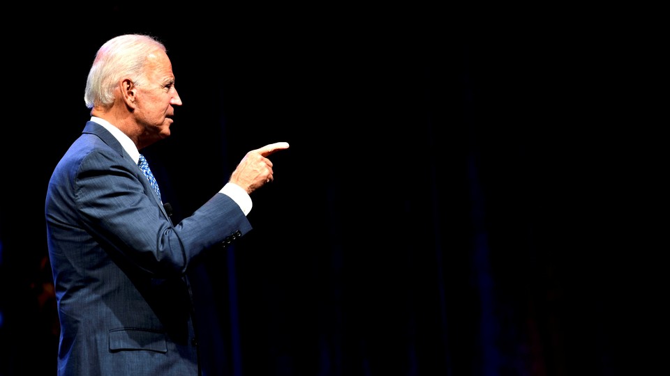 In profile, Joe Biden stands in front of a black background and points in front of him.