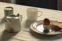 A table covered in a white table cloth, and on top, a tea kettle, two mugs, and a plate with an egg, a spoon, and a slice of bread