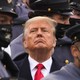 Donald Trump surrounded by uniformed cadets, looking upward with a stern expression.