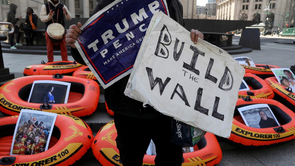 A person holding a "Trump, Pence" support sign and a poster reading "Build the Wall" stands in front of rafts meant to display opposition to the U.S. refugee ban in New York City.