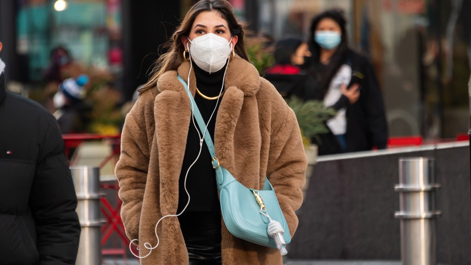 a person wearing a faux fur coat and face mask, with a blue bag over their shoulder