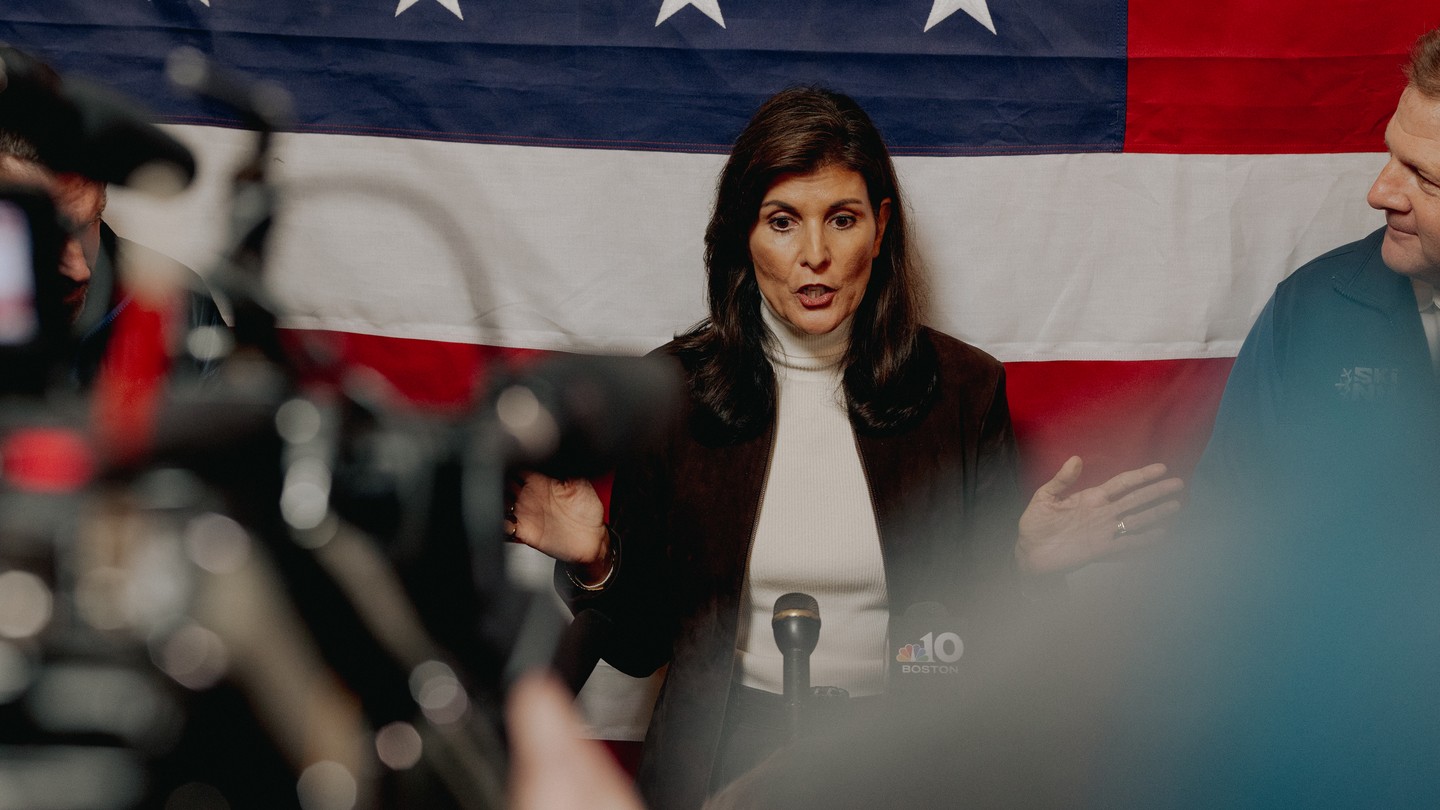 Photo of Nikki Haley speaking at a microphone, hands apart, with press before her and an American flag behind her