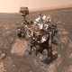 A selfie of a large Mars rover, with a reddish-colored dry, rocky, and dusty Martian landscape in the background.