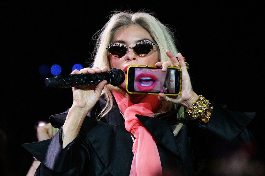 A performer sings onstage while holding a smartphone to their face that is recording video, displaying their mouth on the screen.