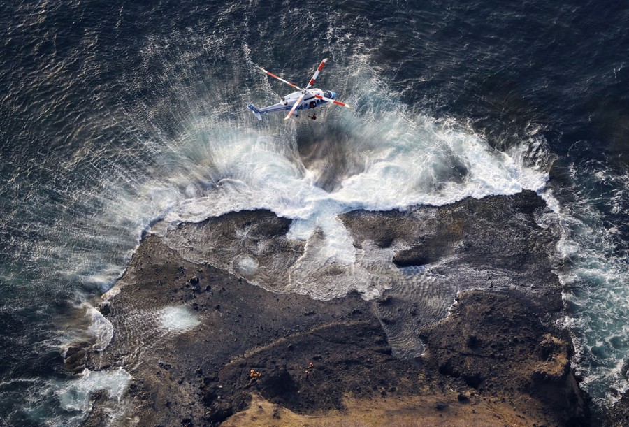 A helicopter hovers low above a rocky shoreline.