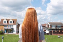 A white girl with long red hair, wearing a gray t-shirt, has her back to the camera and looks out at two lawns and two houses on a suburban street.