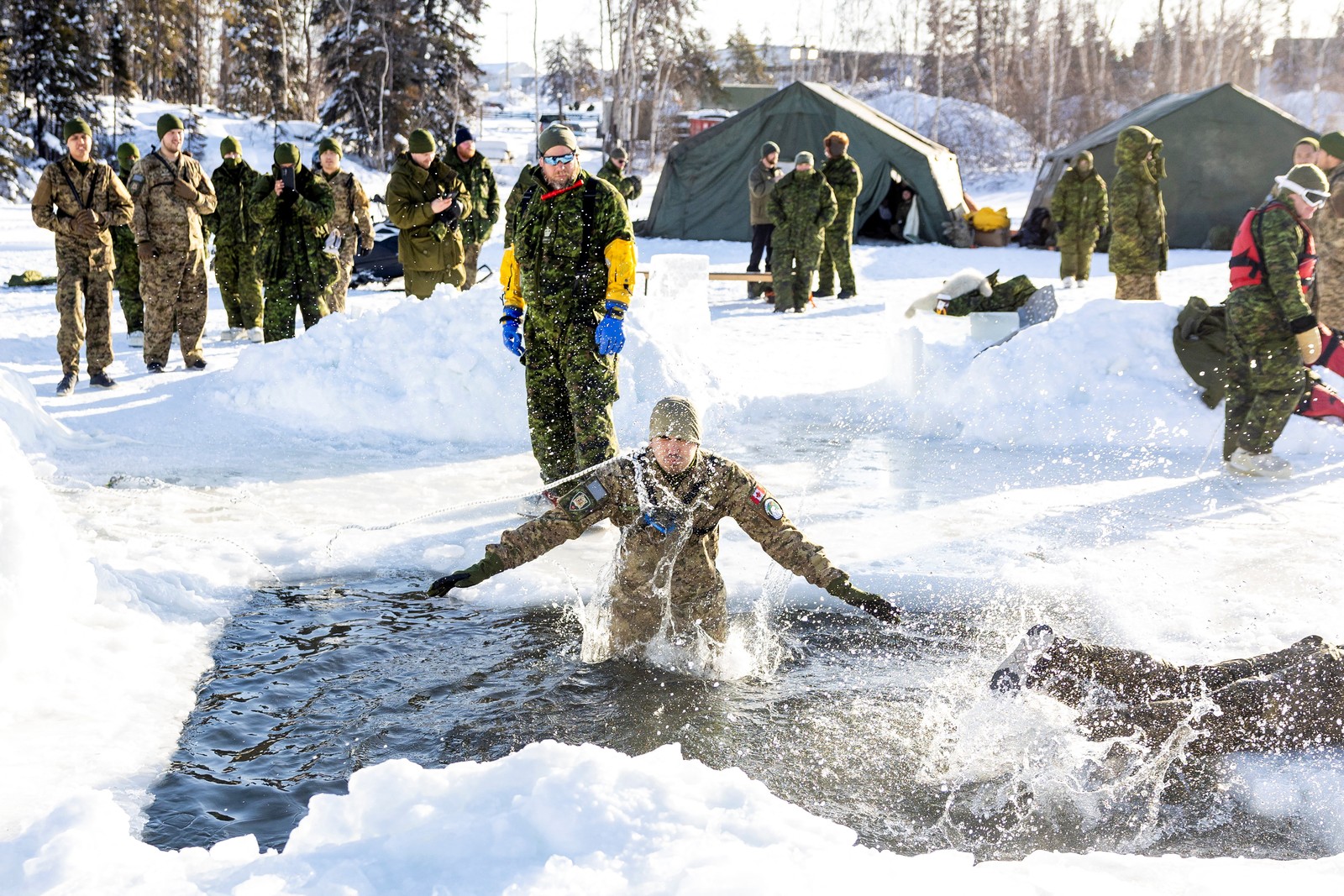 Soldiers stand nearby, watching as another soldier jumps into freezing water, in a hole cut in ice, during a training session.