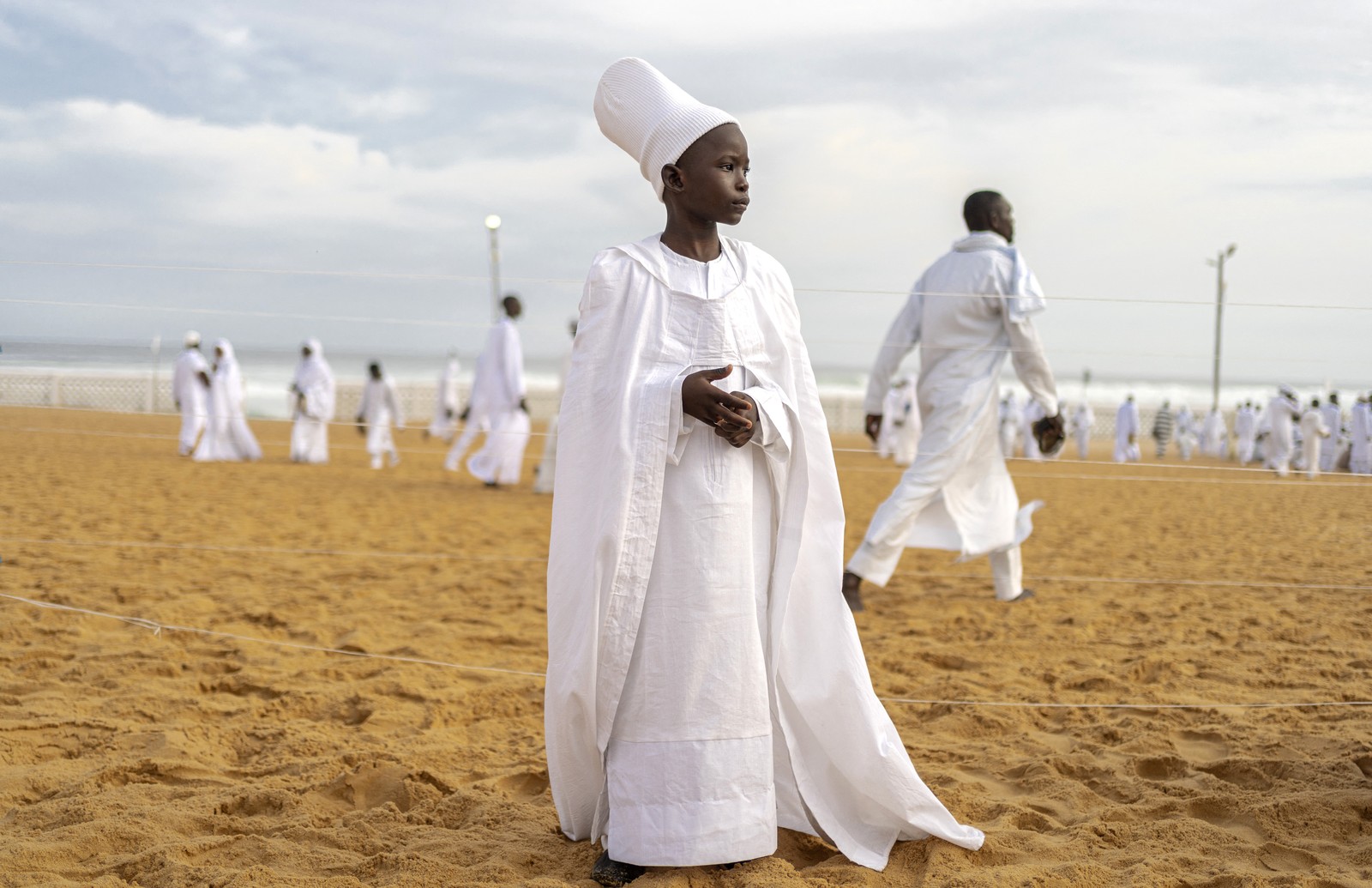 A young person, dressed in a white robe, stands on a beach, with many others dressed in white in the background.