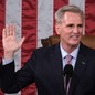 Kevin McCarthy sits in a leather chair at a microphone, holding his right hand aloft. The stripes of the American flag are his backdrop.