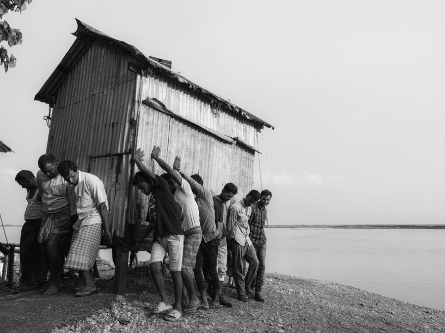 A dozen or so men work together to lift and carry a small shed near a river shoreline.