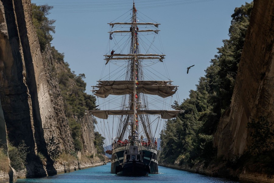 A three-masted sailing ship passes through a narrow canal with tall, steep cliff walls on both sides.