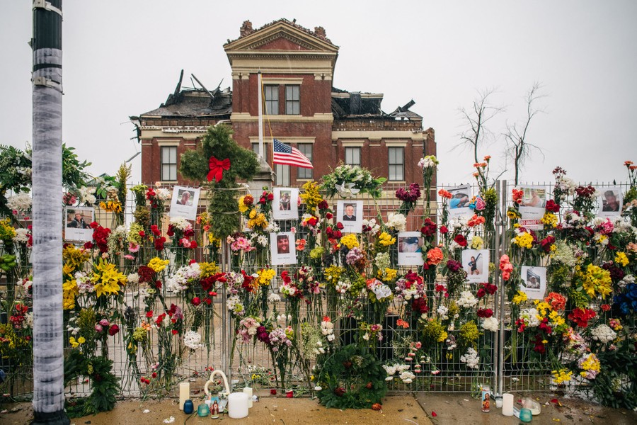 Flower bouquets and portraits cover a temporary fence in front of a damaged courthouse, as a makeshift memorial.