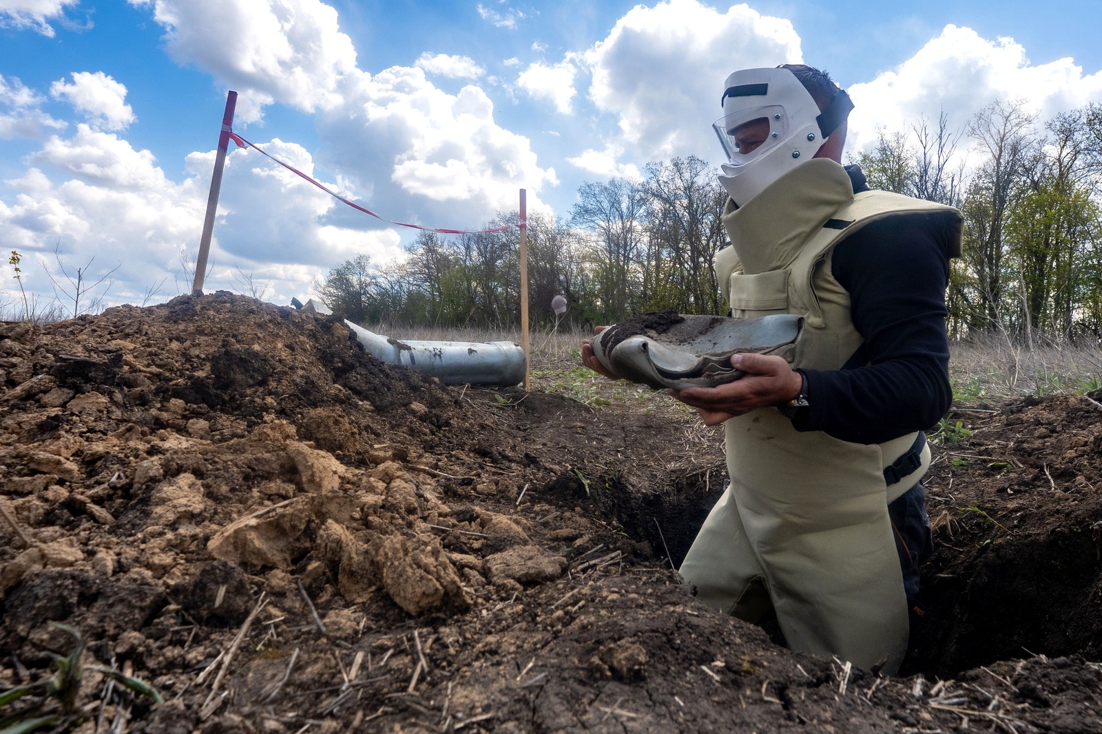 A person wearing protective gear and a face mask removes potential explosive material from a hole in a farm field.