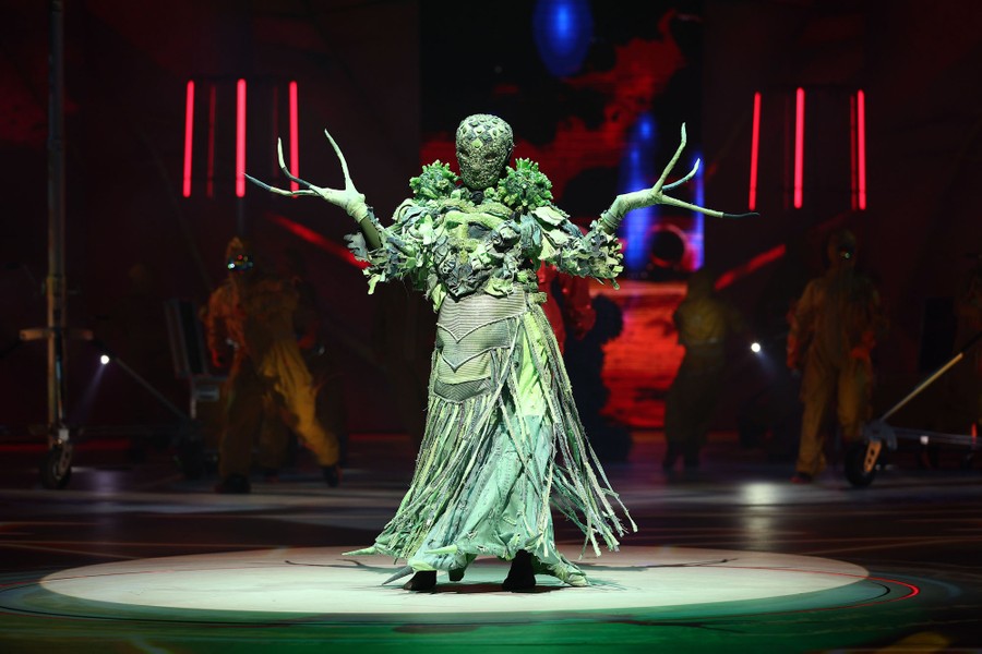 A model displays a layered costume with a full head mask and long finger extensions.