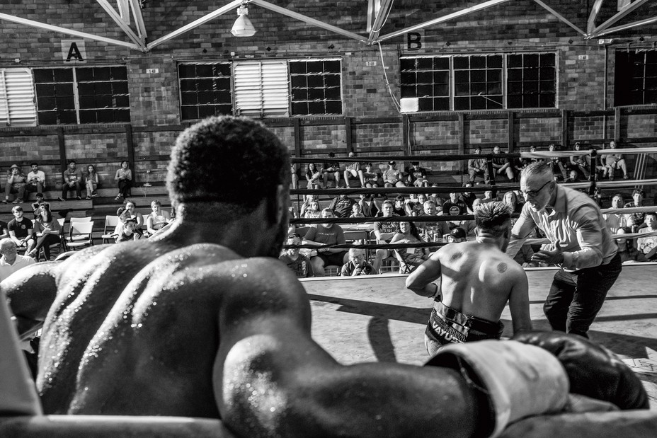 photo taken from corner of ring behind boxer's shoulders and arms, with ref talking to other boxer on his knees in center of ring and audience behind
