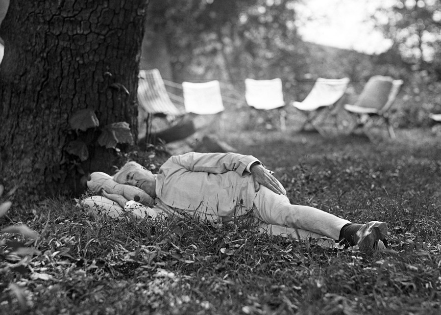 Thomas Edison naps in the grass, his head on pillows, beneath a large tree.