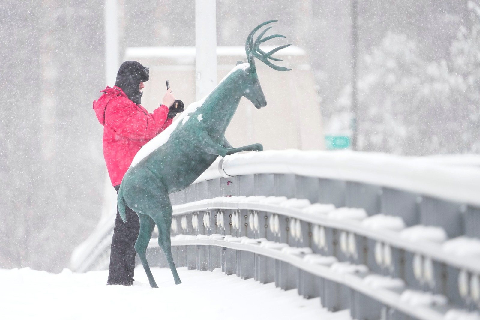 A person takes a photograph from atop a snow-covered bridge, standing beside a statue of a deer leaning on the railing.