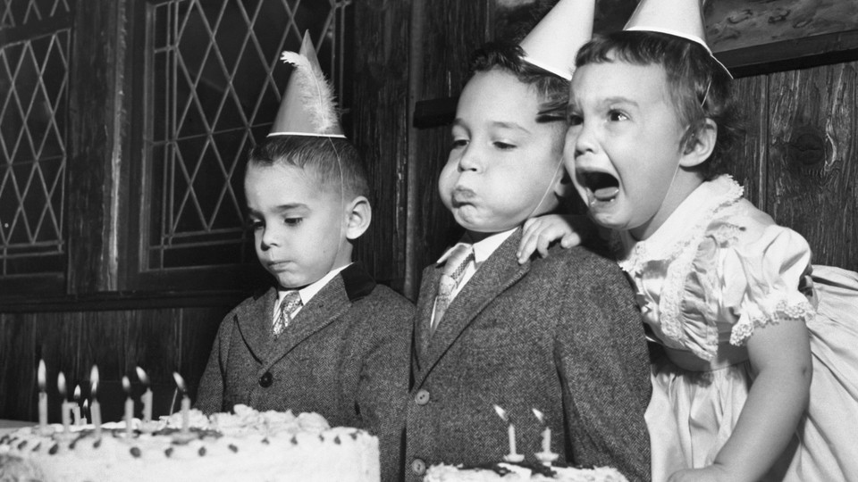 Three siblings with their birthday cake. One appears indifferent, one is blowing out the candles, one is crying.