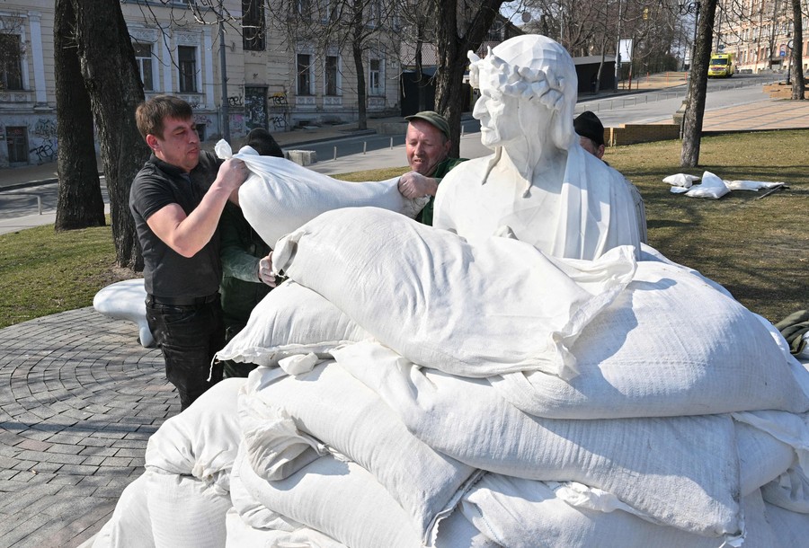 Two people lift sandbags onto a pile surrounding a statue on a city street.