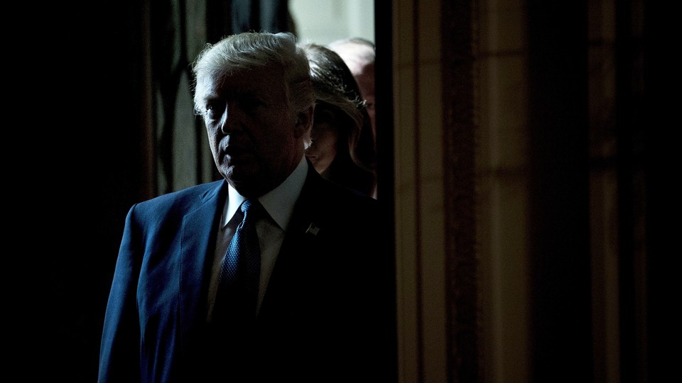 A 2017 photo of then-President Donald Trump wearing a blue suit and tie, standing in a dark corridor