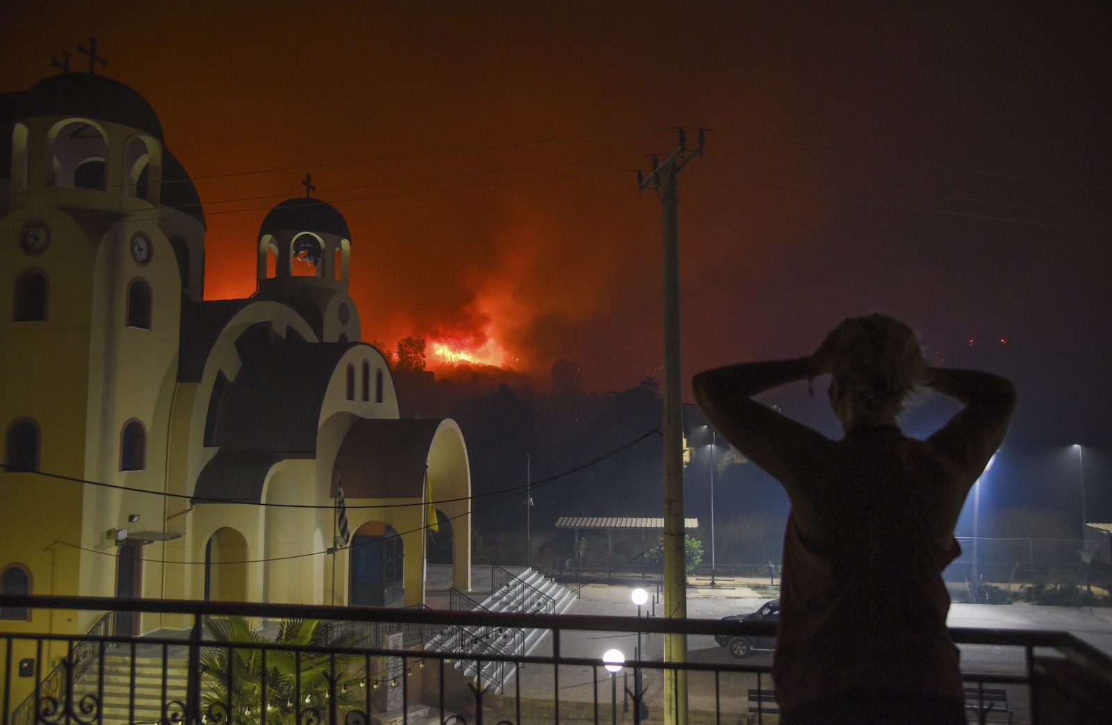 A person reacts, hands on their head, looking out toward a wildfire on a nearby hillside.