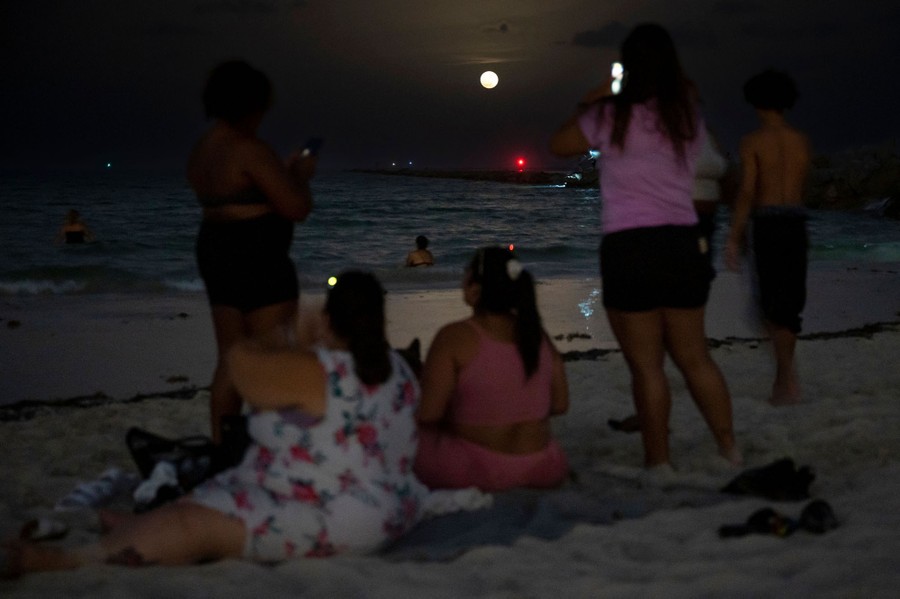 About nine people sit, stand, and swim on a beach at moonrise.