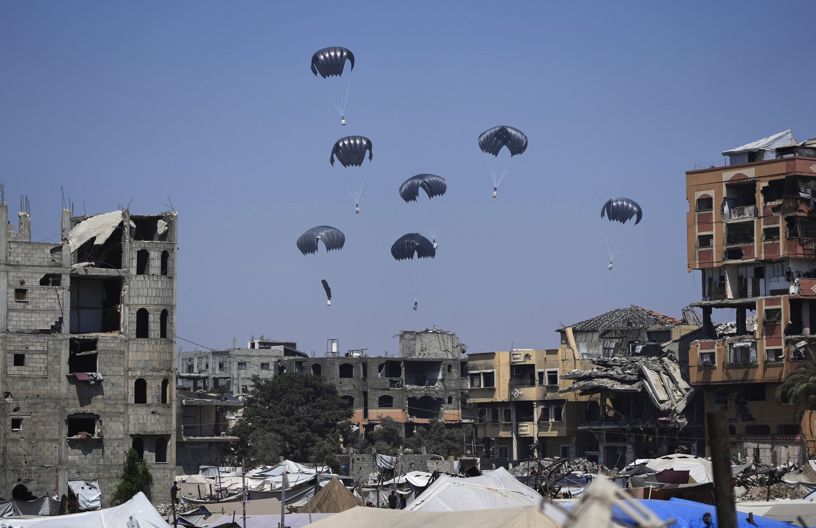 Crates of food and aid drop from the sky by parachute, seen over ruined structures in Gaza.