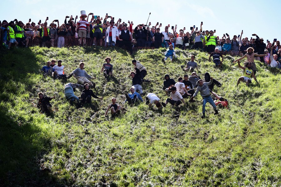 Several dozen racers run, jump, tumble, and fall down a steep, grass-covered hill.