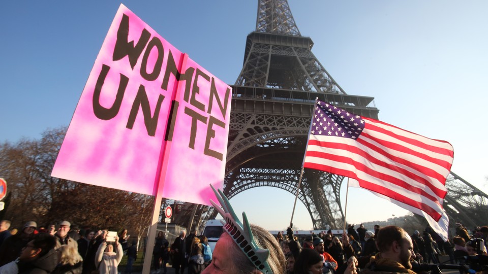 Protesters attend the Women's March in Paris in 2017. One holds an American flag and one holds a sign that reads "Women unite."