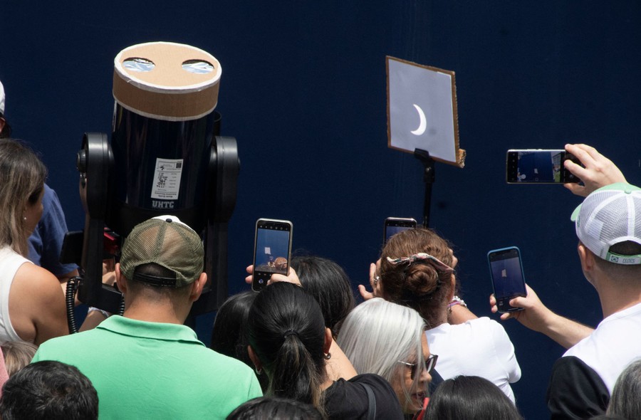 A small crowd looks at and takes pictures of the image of a partial solar eclipse, visible on a board beside a large telescope.