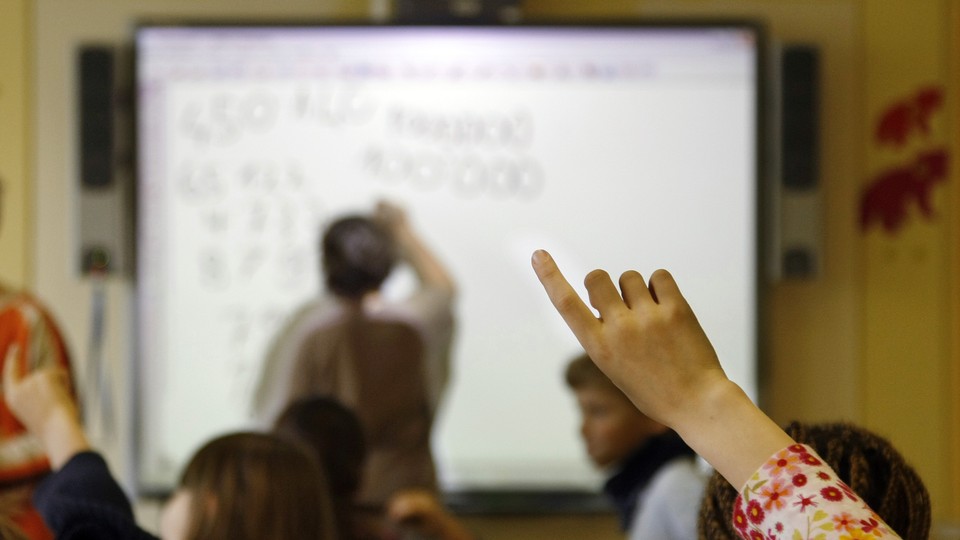 A student raises her hand as her teacher leads a lesson on a SmartBoard. Questions are being raised about whether teachers should be paid for the lessons they create.