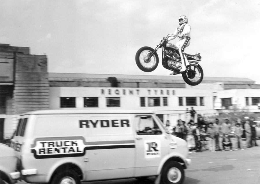 Evel Knievel, seen in mid-air, on a motorcycle, during a jump over several rental vans.